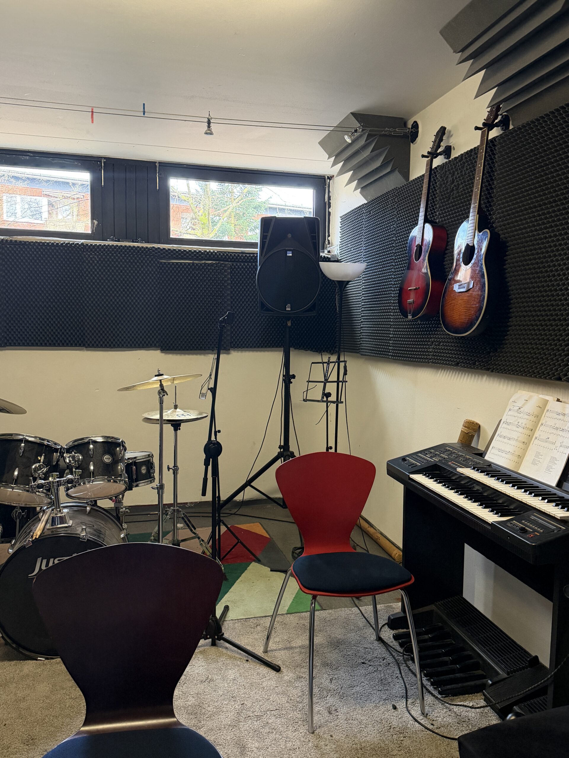 Proberaum mit Schlagzeug, E-Piano und zwei an der Wand hängenden Gitarren. Die Wände sind mit schwarzem Schallschutzschaum verkleidet, Lautsprecher und Mikrofonständer stehen bereit.