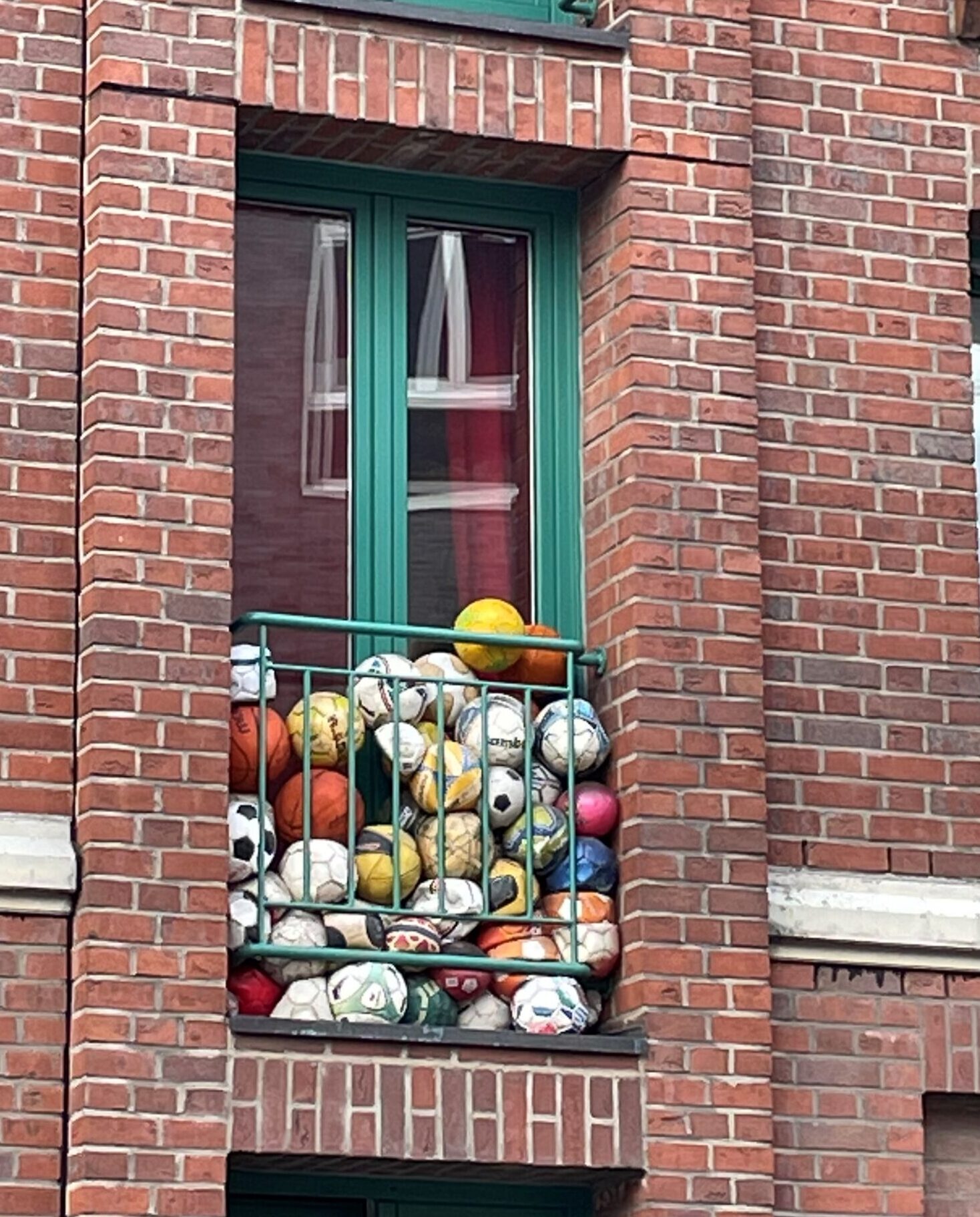 Mehrstöckiges Wohnhaus mit roter Backsteinfassade und grünen Fensterrahmen. Auf einem kleinen Balkon im ersten Stock sind zahlreiche bunte Sportbälle – vor allem Fußbälle, aber auch andere Ballarten – dicht gestapelt und füllen den Balkon bis zum Geländer.