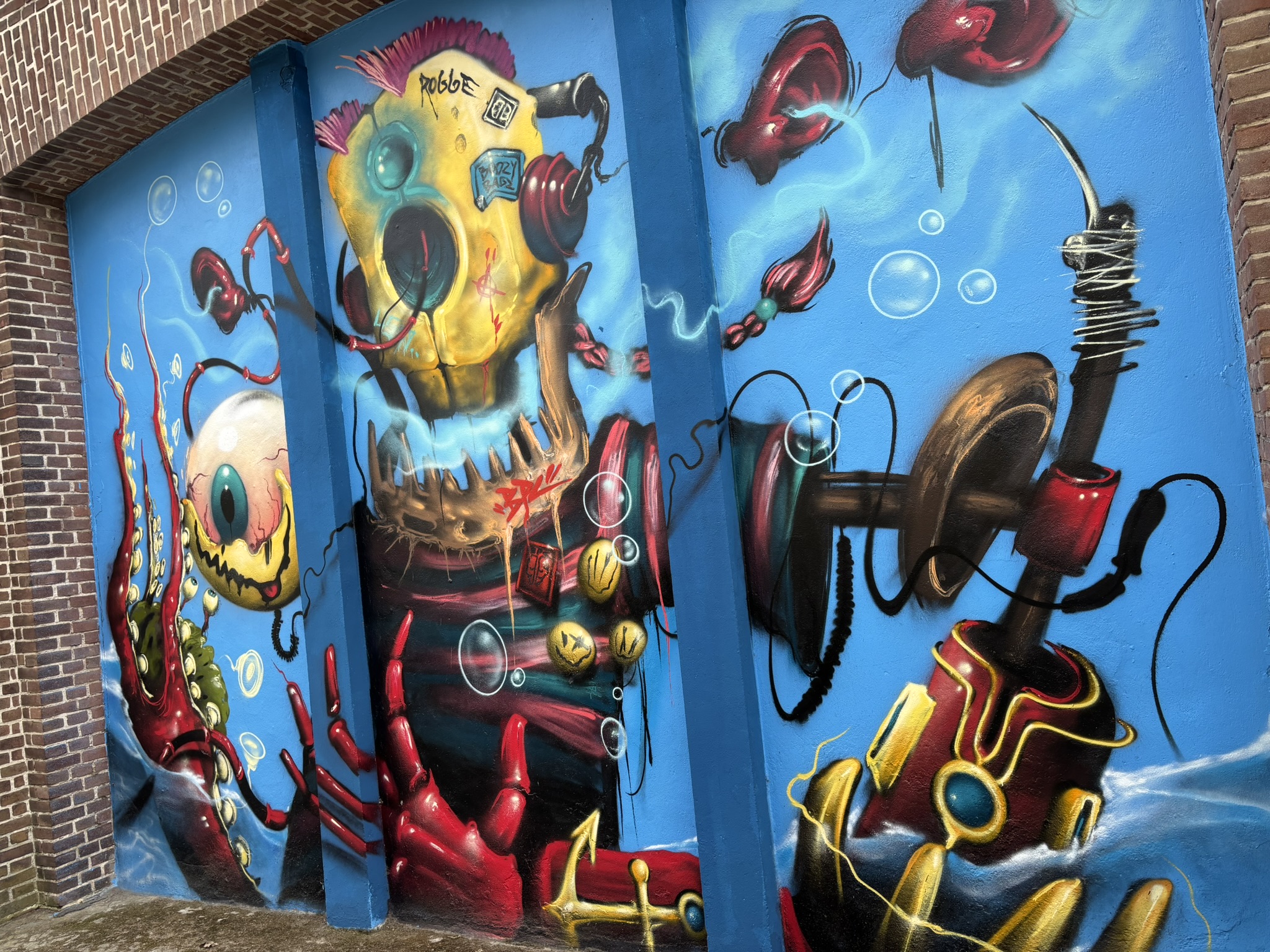 Buntes Graffiti mit fantasievollen Unterwasserwesen vor blauem Hintergrund. Zu sehen sind ein großes Monster mit Kopfhörern, ein einäugiges Wesen, Luftblasen und mechanische Elemente in Rot, Gelb und Gold.